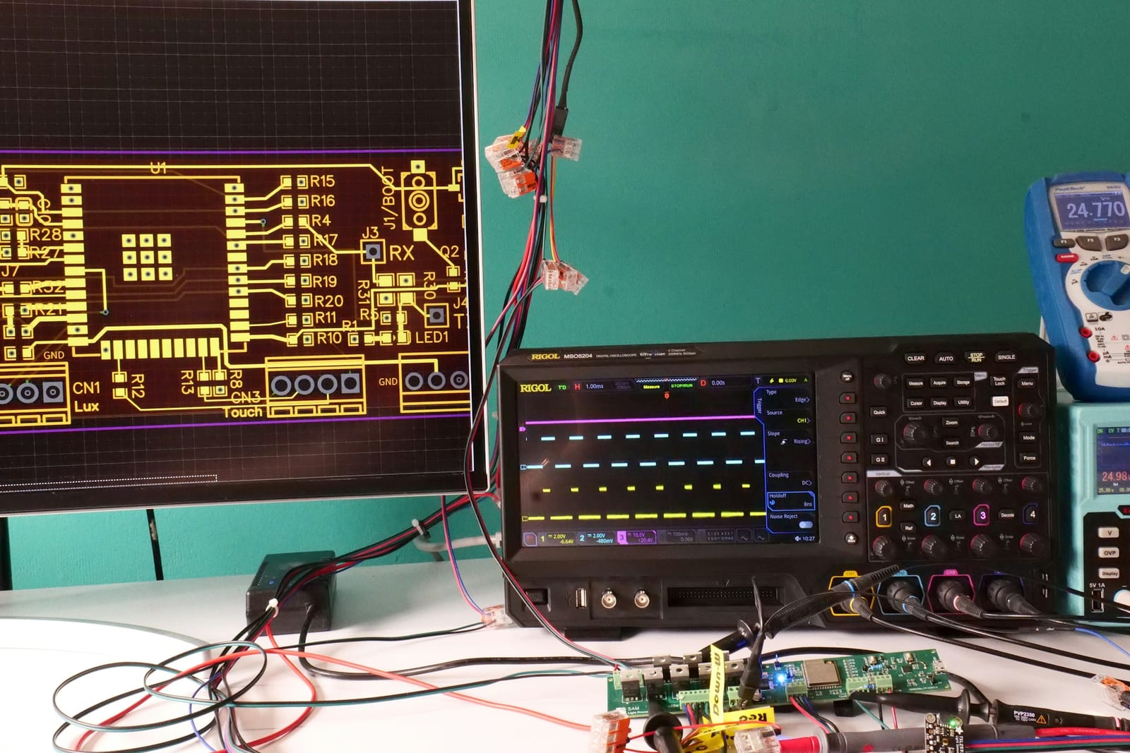 工程師的工作檯：螢幕上是 PCB 佈局圖，中央一台示波器，前景是接滿探棒線的開發板